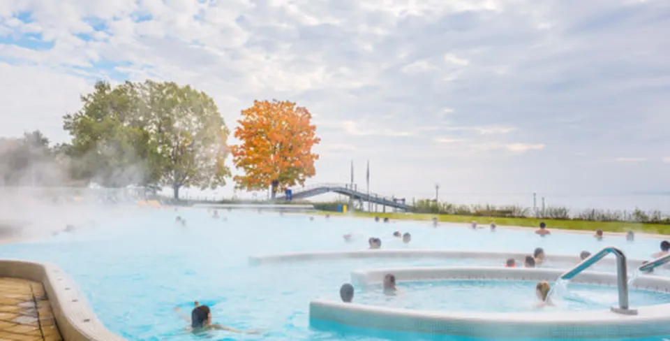Menschen entspannen in einem dampfenden Außenpool. Bäume mit Herbstlaub im Hintergrund und ein bewölkter Himmel.