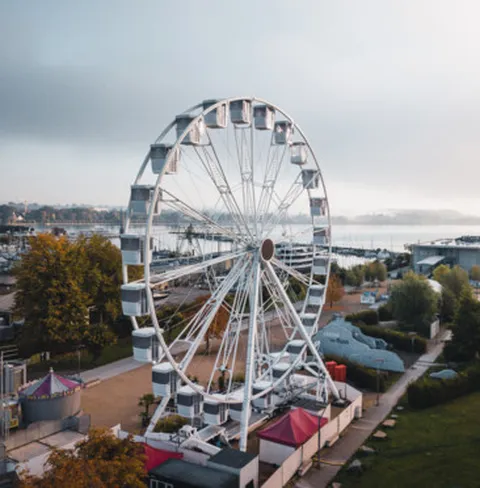 Ein großes Riesenrad steht am Ufer eines Sees, umgeben von Bäumen und Gebäuden, bei leicht bewölktem Himmel.