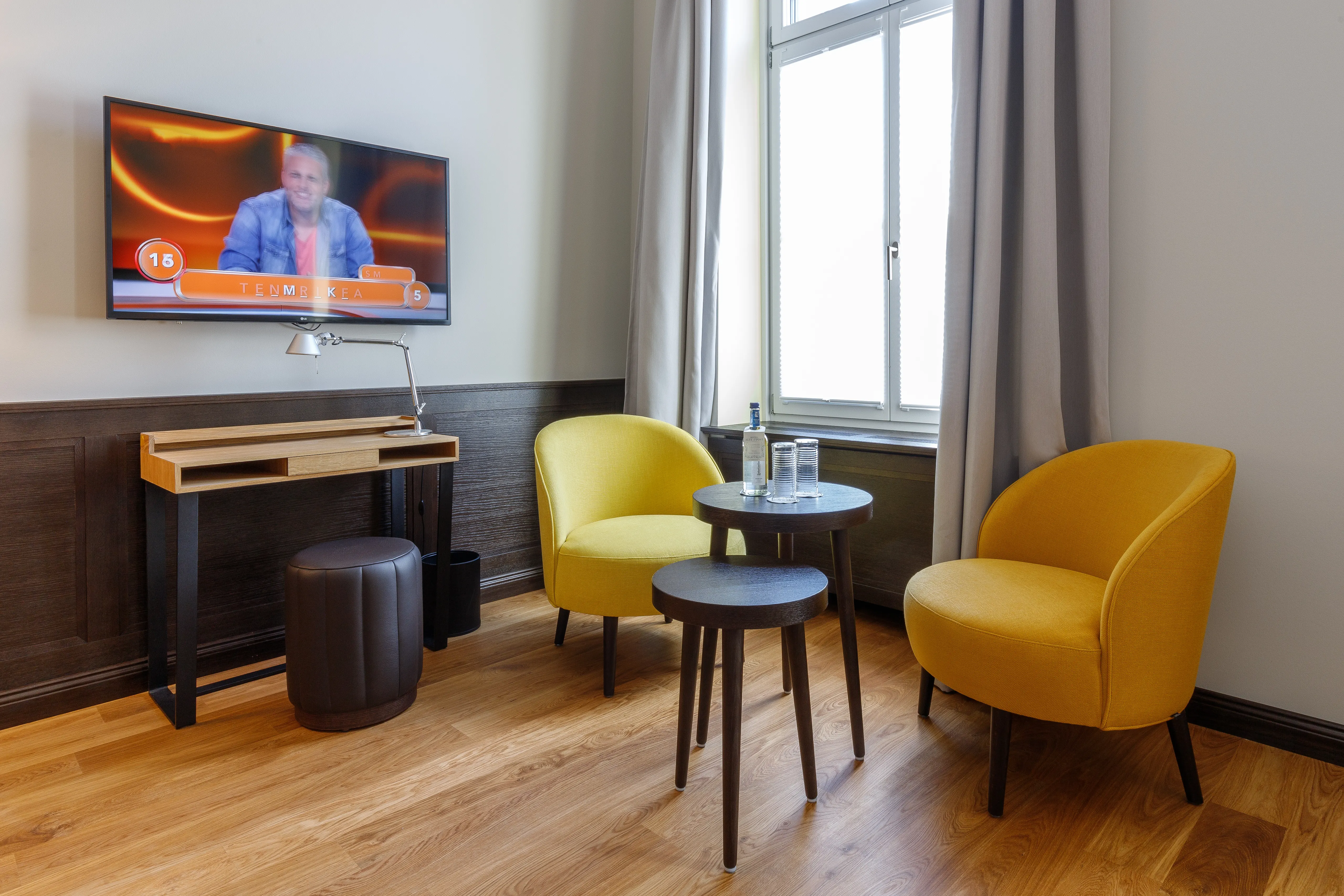 Ein moderner Raum mit zwei gelben Sesseln, einem kleinen Tisch, Flaschen und Gläsern, einem Flachbildfernseher und einem Schreibtisch.