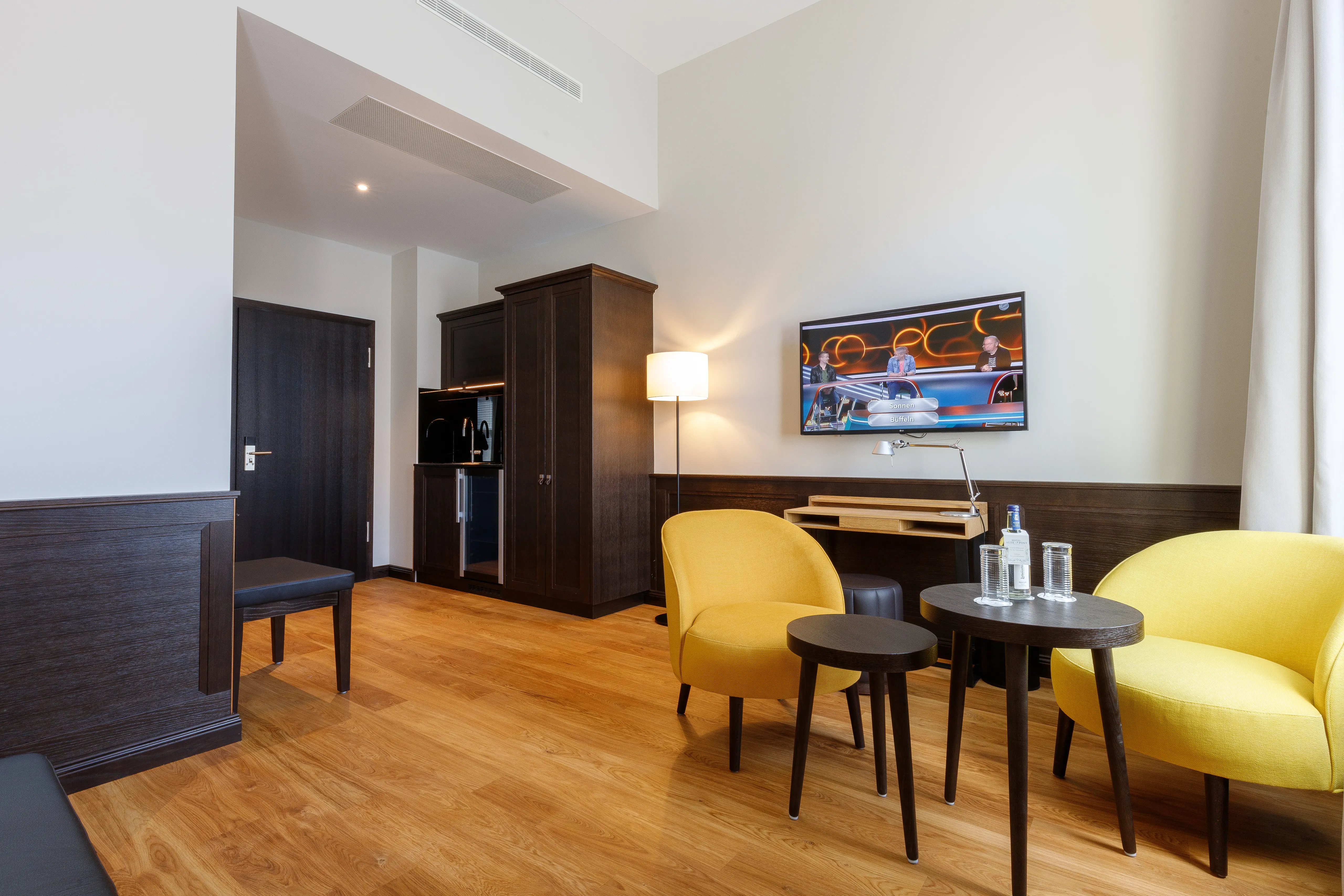 Moderner Raum mit Holzboden, zwei gelben Sesseln, kleinem Tisch, Flachbildfernseher an der Wand und Kochnische im Hintergrund.