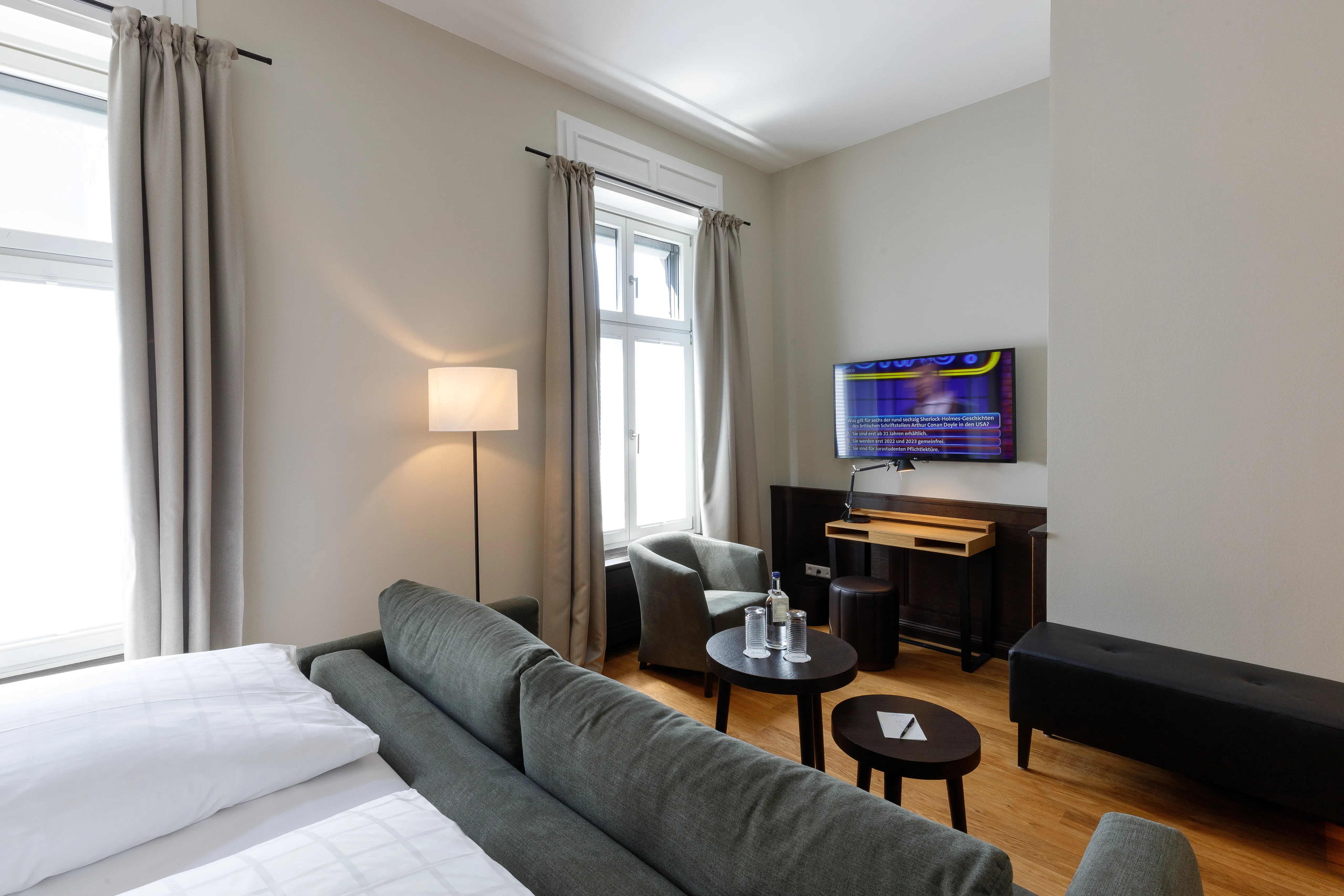 Modernes Hotelzimmer mit grauem Sofa, Tisch mit Wasserflaschen, Fernseher an der Wand, Schreibtisch, Stuhl, Stehlampe und großen Fenstern mit Vorhängen.