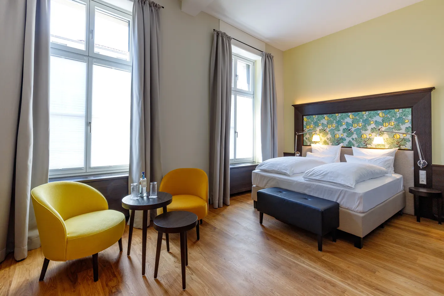 Helles Hotelzimmer mit Doppelbett, buntem Kopfteil, zwei gelben Stühlen, zwei Tischen und großen Fenstern mit Vorhängen.
