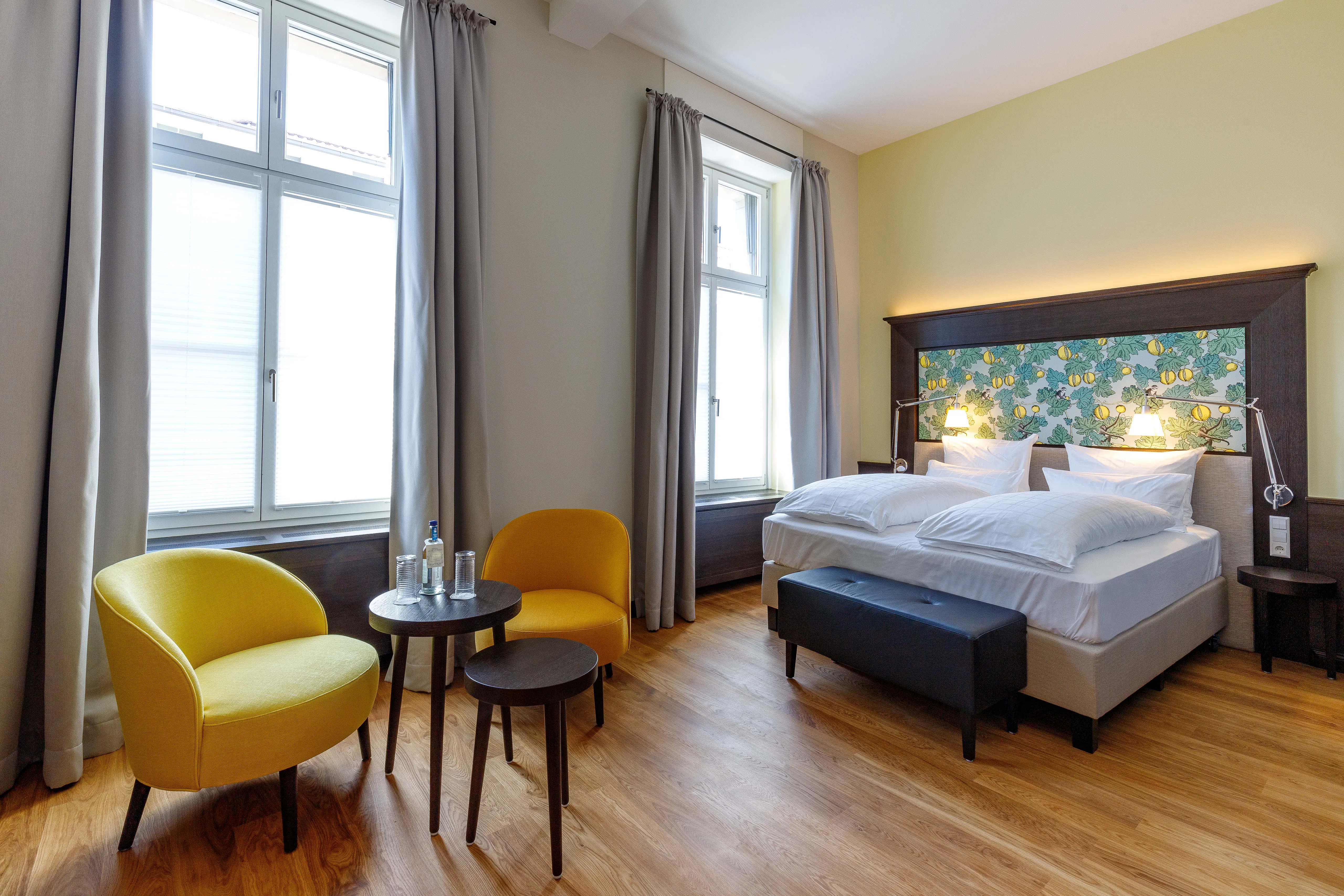 Helles Hotelzimmer mit Doppelbett, buntem Kopfteil, zwei gelben Stühlen, zwei Tischen und großen Fenstern mit Vorhängen.