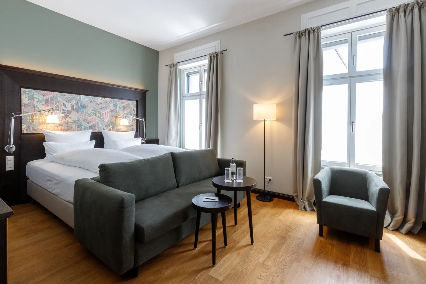 Helles Hotelzimmer mit Doppelbett, grauem Sofa, Sesseln, Beistelltischen und Stehlampe. Zwei große Fenster mit Vorhängen lassen Licht herein.