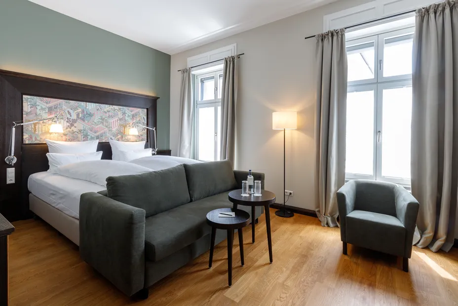Helles Hotelzimmer mit Doppelbett, grauem Sofa, Sesseln, Beistelltischen und Stehlampe. Zwei große Fenster mit Vorhängen lassen Licht herein.