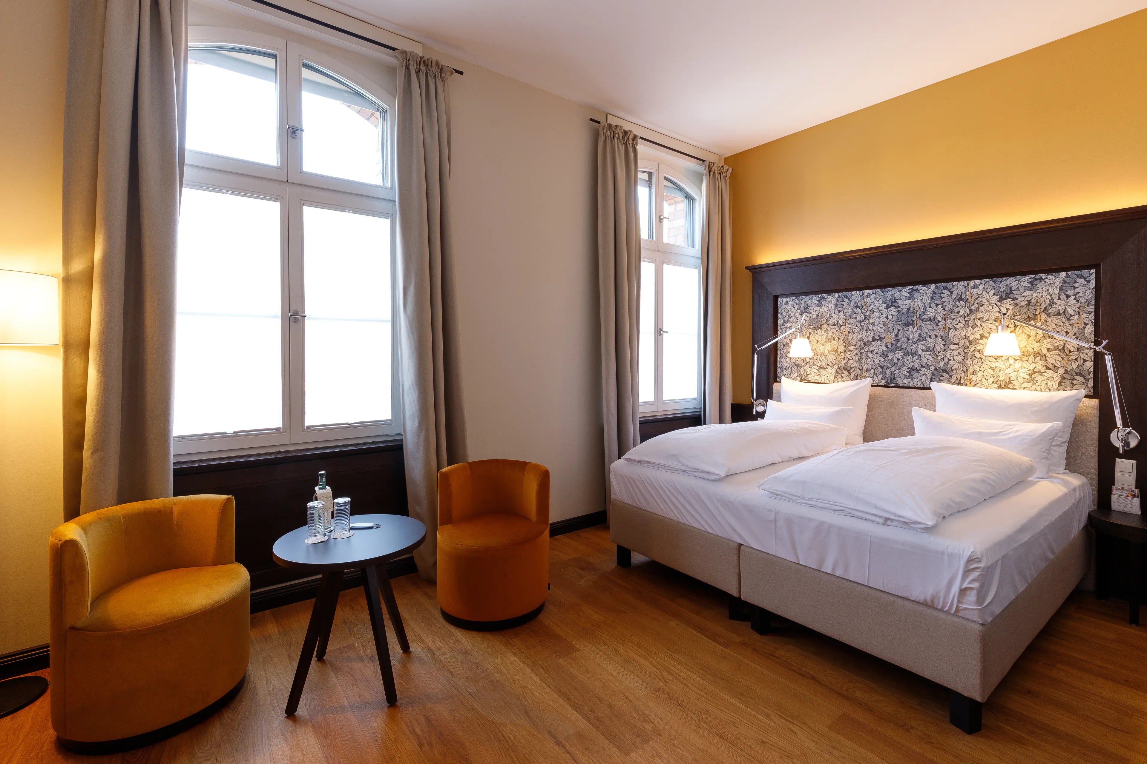 Gemütliches Hotelzimmer mit Doppelbett, zwei gelben Sesseln, Tisch, zwei Fenstern und warmem Licht, Holzboden und gelber Wand.