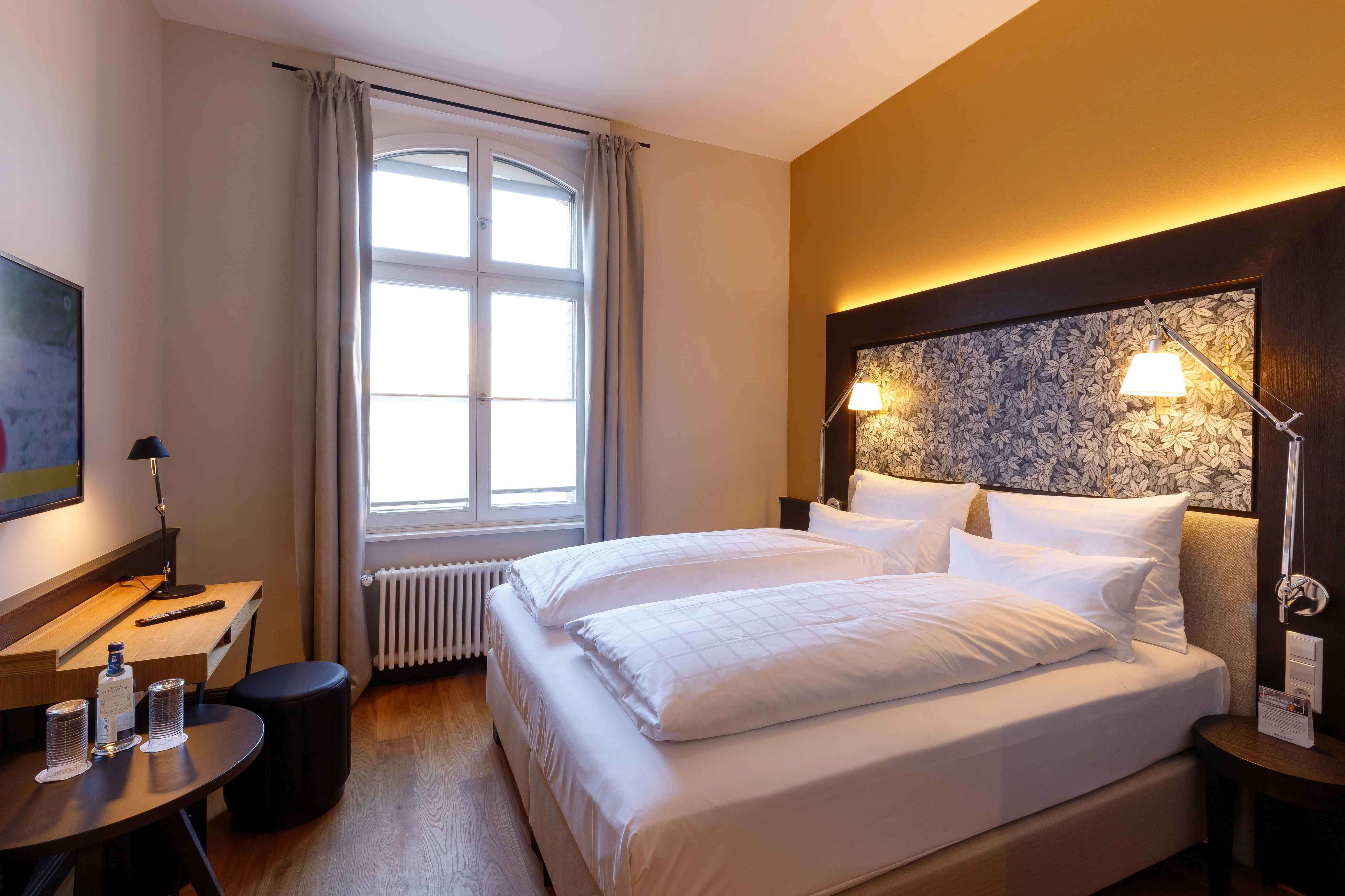 Gemütliches Hotelzimmer mit Doppelbett, dekorativer Wand, Schreibtisch, Fernseher und Fenster mit Tageslicht.