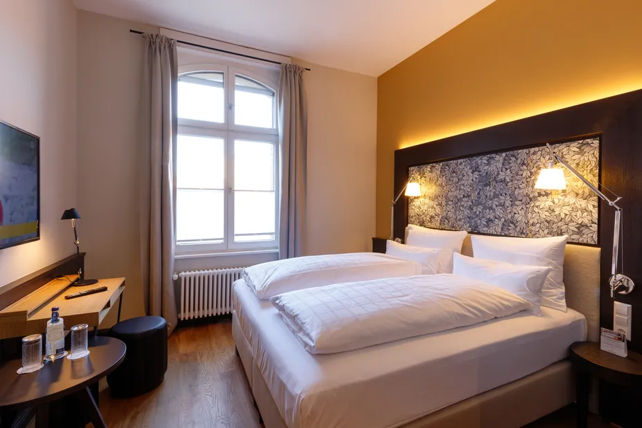 Gemütliches Hotelzimmer mit Doppelbett, dekorativer Wand, Schreibtisch, Fernseher und Fenster mit Tageslicht.