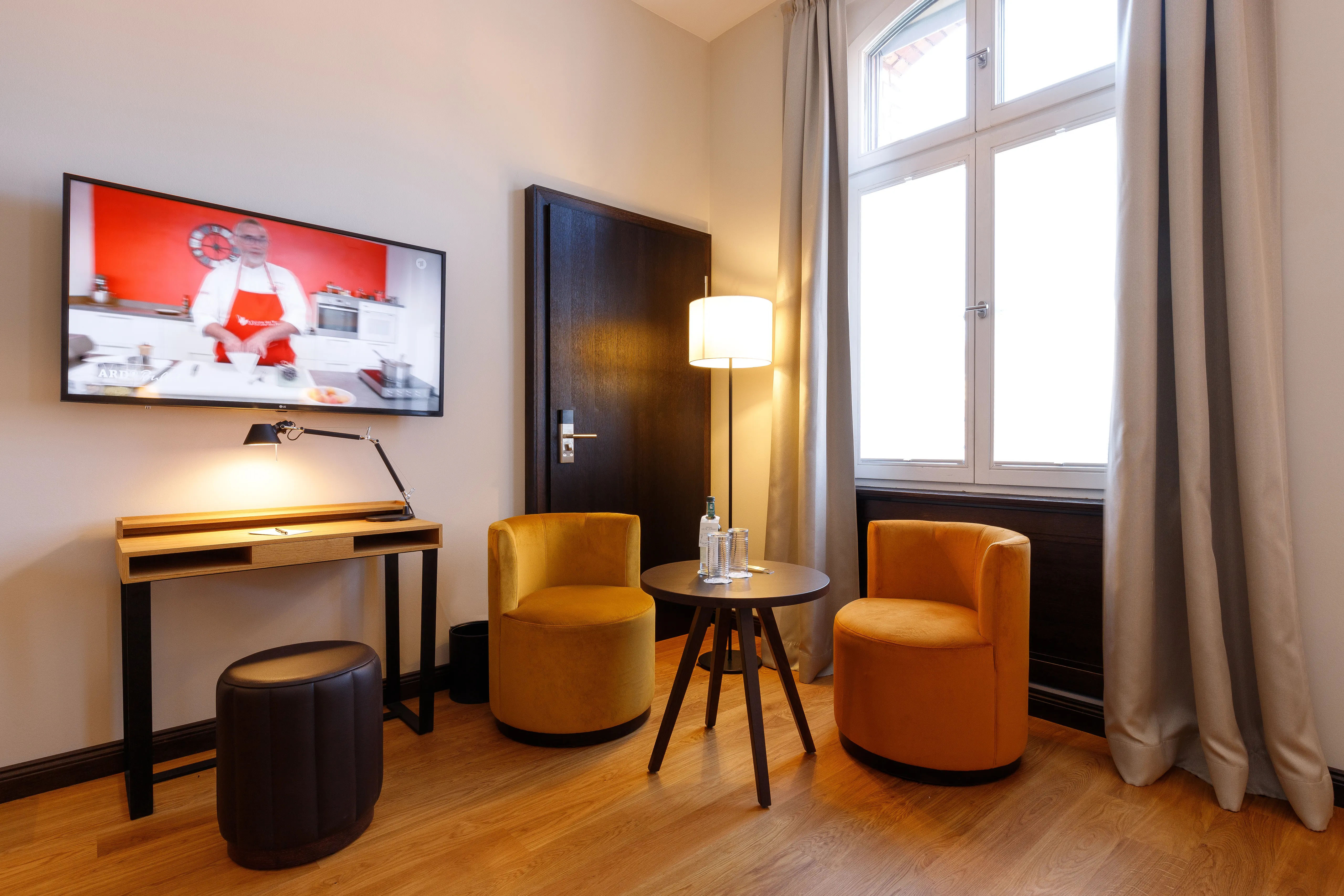 Gemütlicher Raum mit Fernseher, Schreibtisch, zwei gelben Sesseln, kleinem Tisch mit Wasserflaschen, Stehlampe und großem Fenster mit Vorhängen.