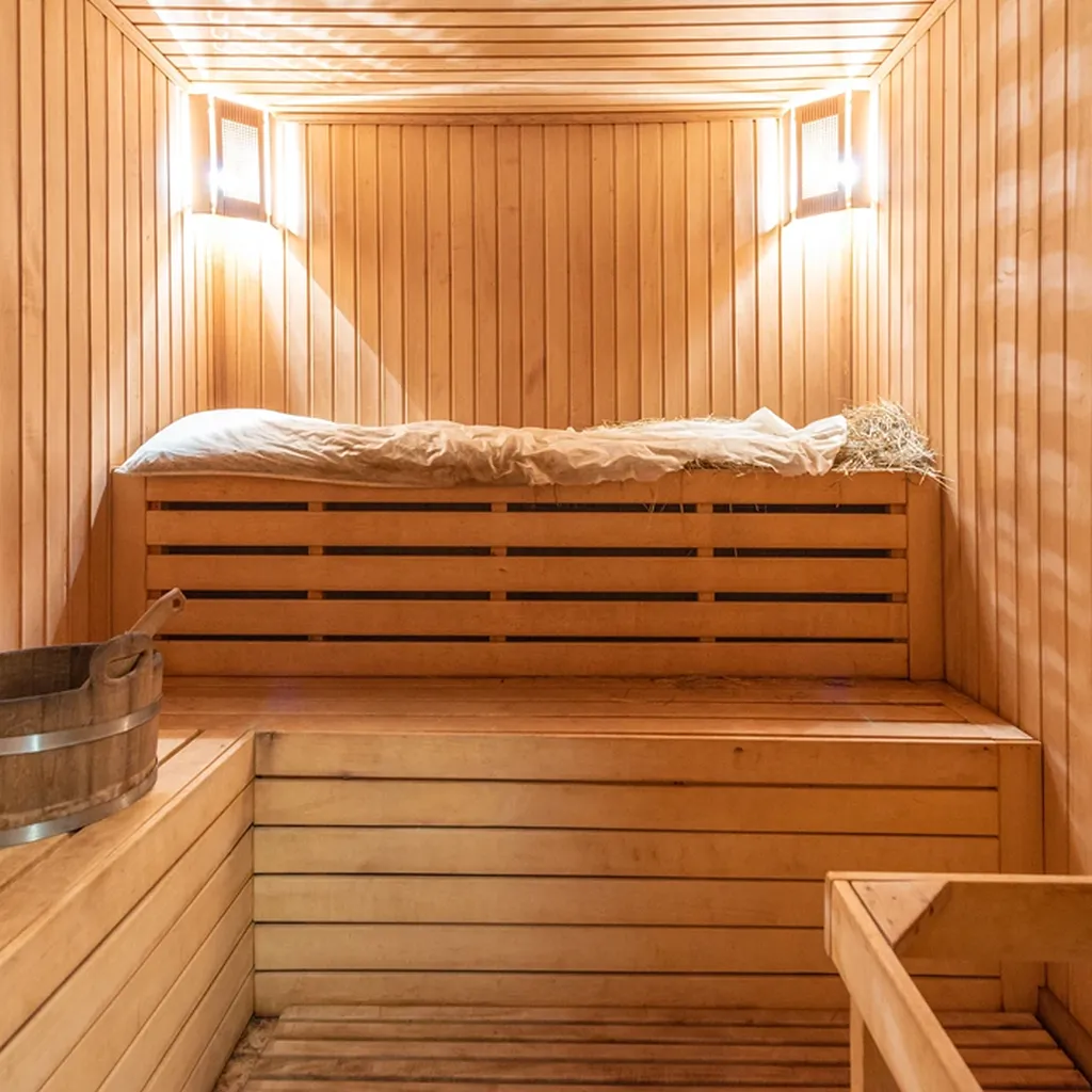 Innenraum einer Sauna mit Holzvertäfelung, einer Liegefläche mit Tuch und Eimer aus Holz, beleuchtet durch warme Lichter an den Wänden.