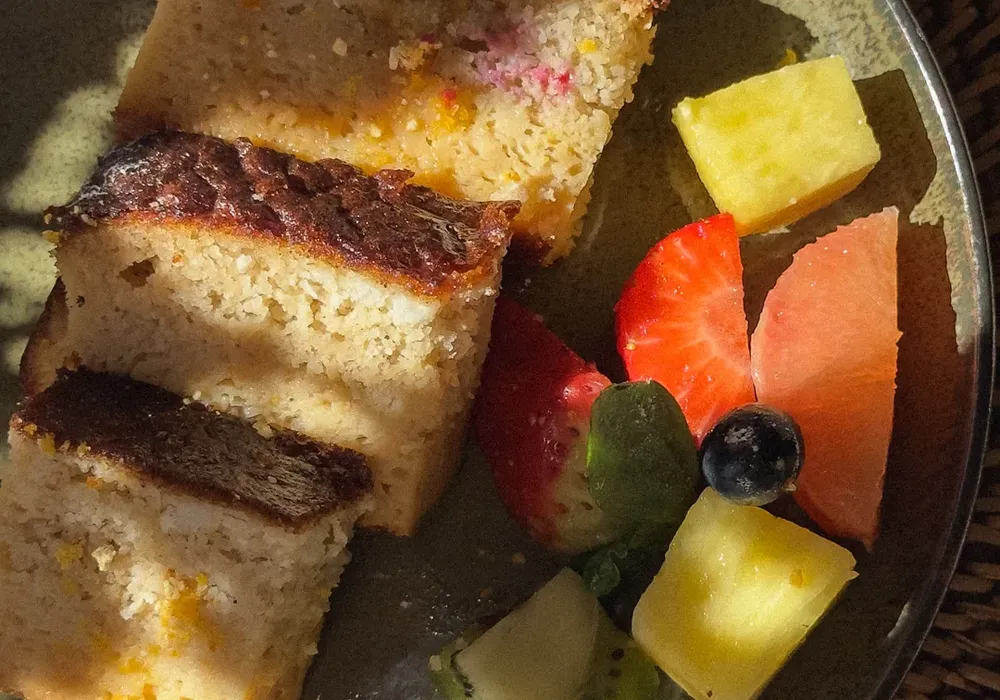 Slices of cake with fresh fruit, including pineapple, strawberry, grapefruit, kiwi, and blueberry, on a green plate in sunlight.