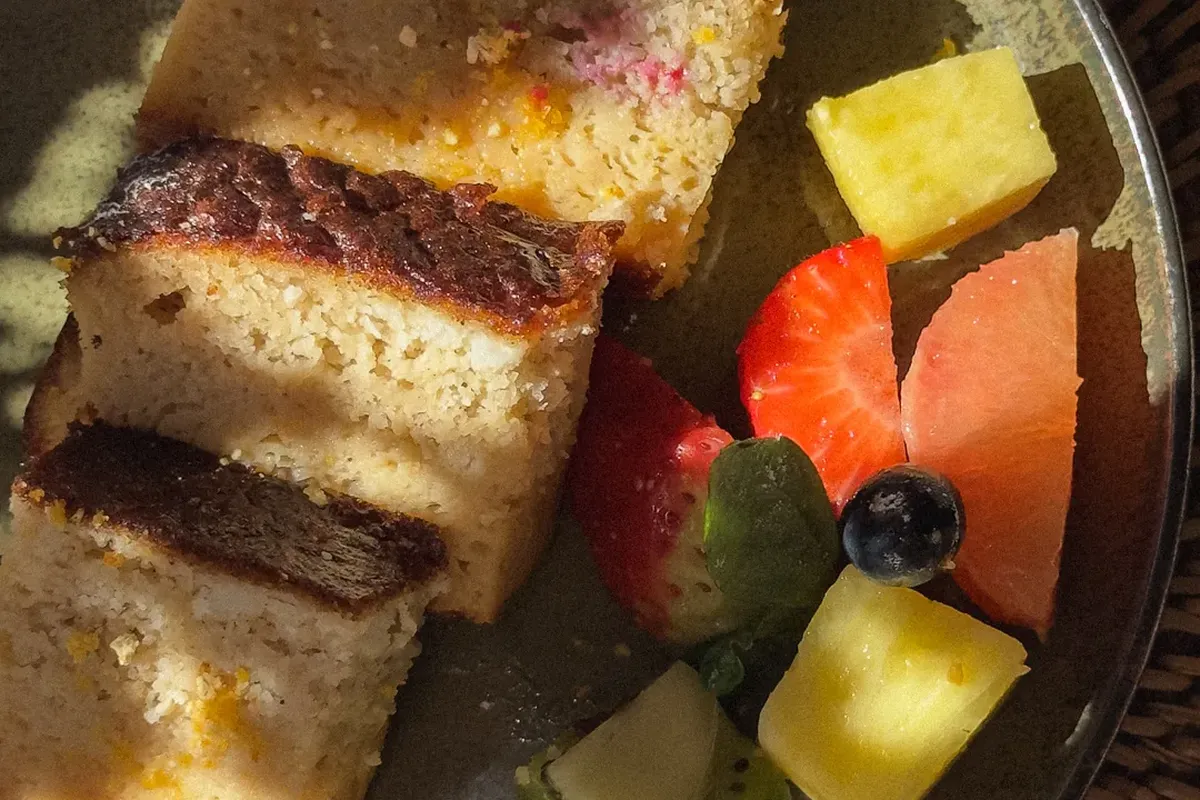 Slices of cake with golden crust next to assorted fruit pieces like pineapple, kiwi, watermelon, and berries on a green plate.