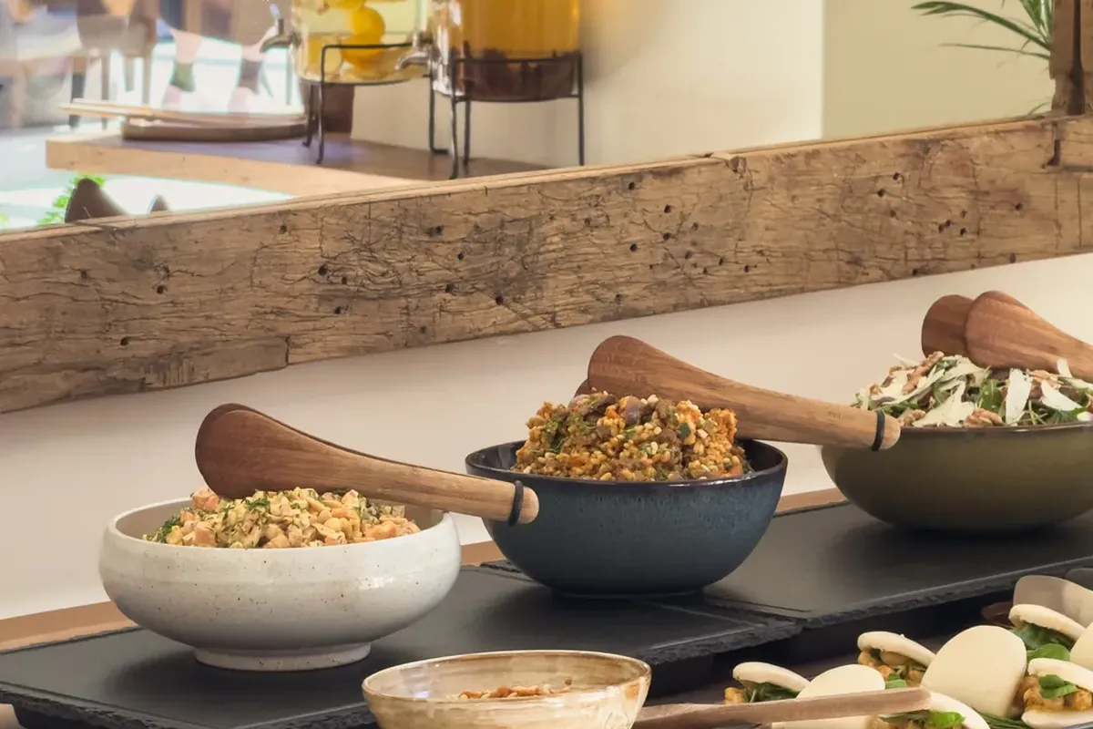 A buffet spread with various bowls of salads, steamed buns, and small appetizers on a wooden counter, with juice dispensers in the background.
