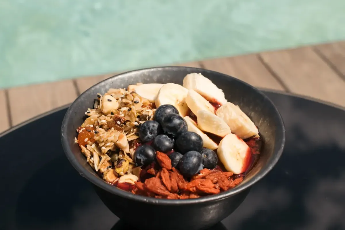 A bowl of granola, bananas, blueberries, and goji berries on a black surface by a pool.
