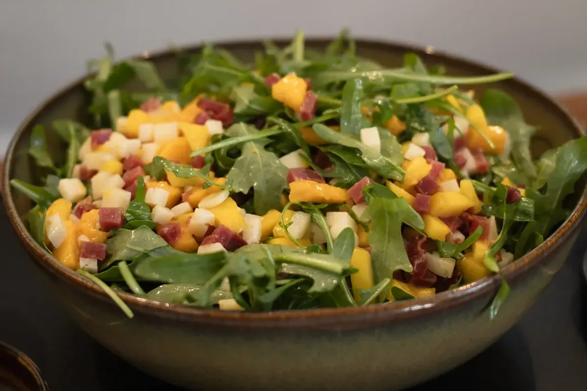 Bowl of mixed salad with arugula, diced cheese, mango, and meat pieces on a dark surface.