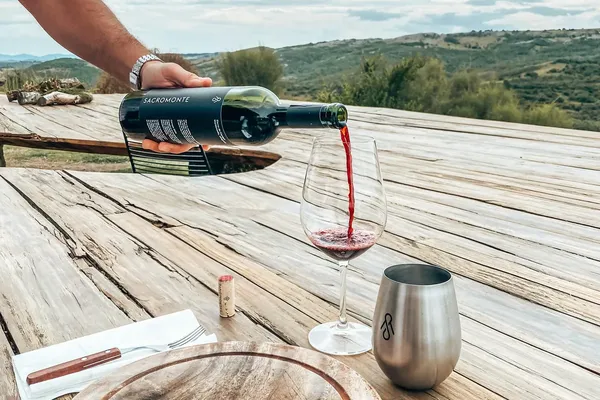 Wine being poured into a glass on a rustic wooden table with a cutting board, knife, and metal cup, overlooking scenic hills.