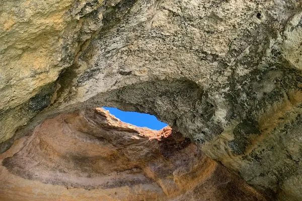 Sunlit interior of a rocky cave with textured walls, featuring a natural circular opening revealing a patch of blue sky above.