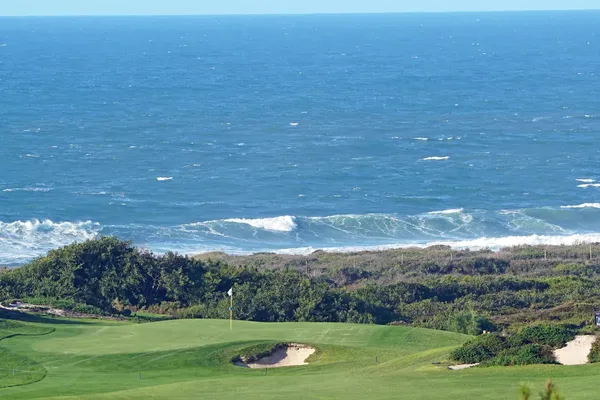 A scenic view of a coastal golf course with lush greens and sand bunkers, bordered by the ocean and waves under a clear blue sky.