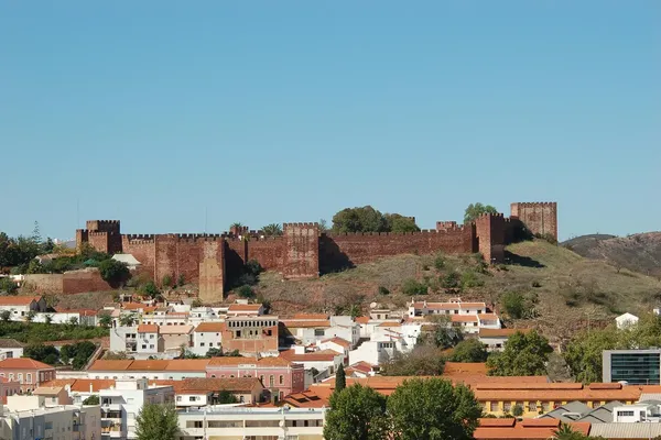 Historic stone castle atop a hill with a town of white buildings and red roofs in the foreground, under a clear blue sky.