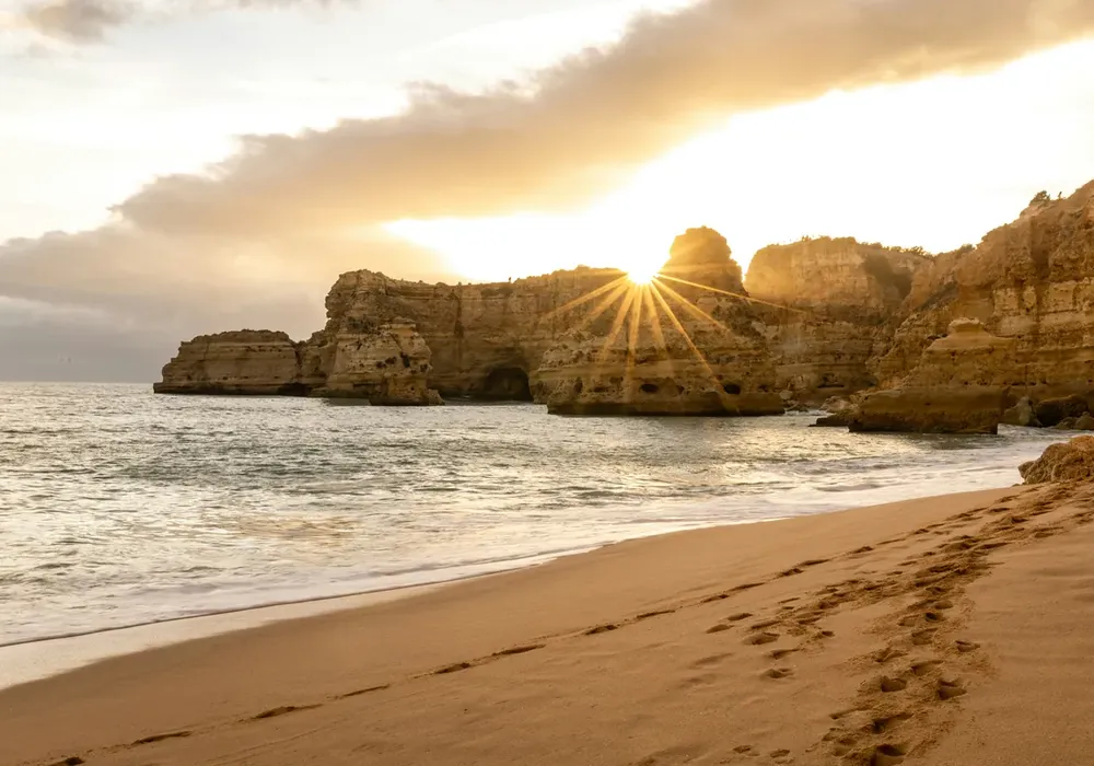 Sunset over rocky cliffs by the ocean, golden sand with footprints in the foreground, and a dramatic sky with clouds.