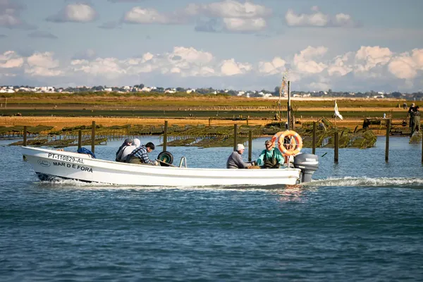 A small motorboat with four people travels on a calm river, passing alongside oyster farms under a partly cloudy sky.