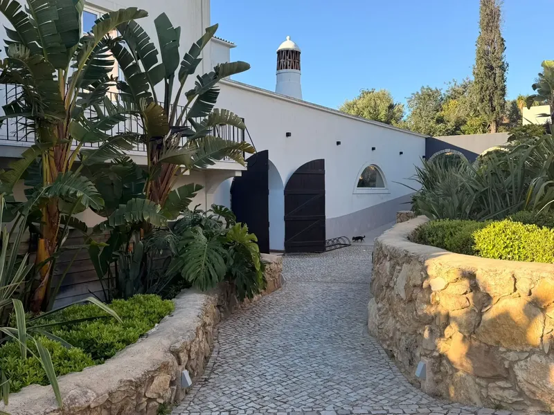 Cobblestone path between white buildings with lush plants and stone walls, leading to a closed gate under a clear blue sky.