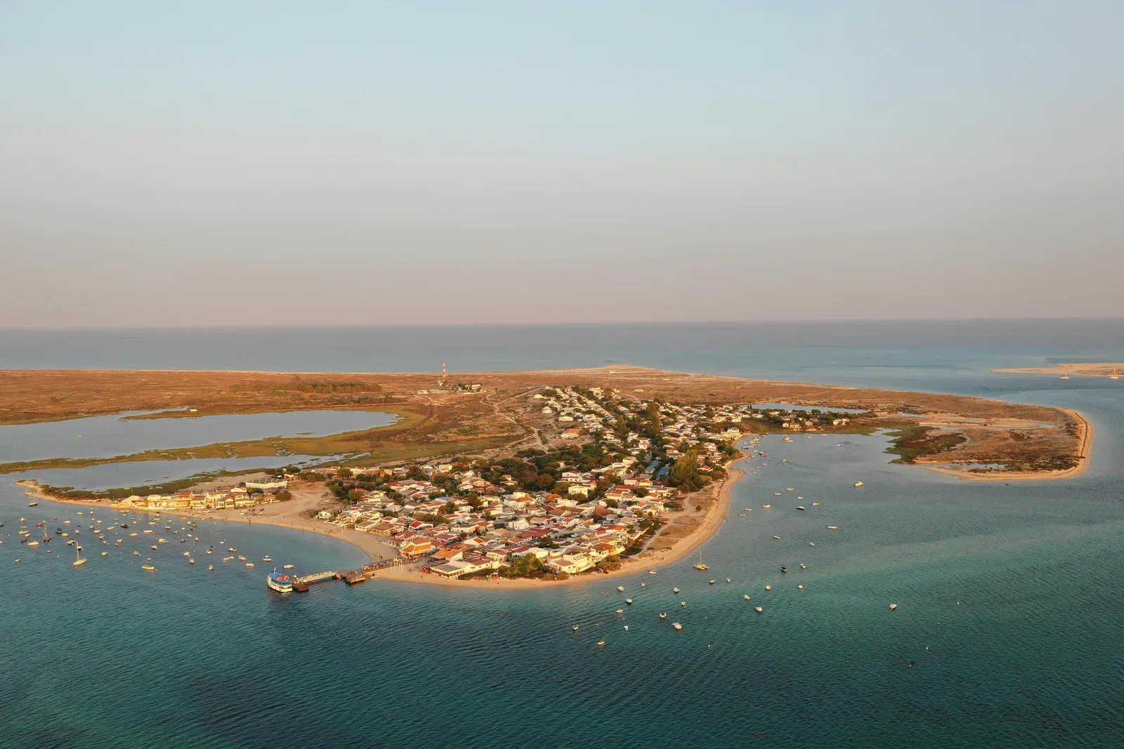 Aerial view of a small coastal town on a peninsula, surrounded by blue water and anchored boats, with a sandy beach and sparse vegetation.