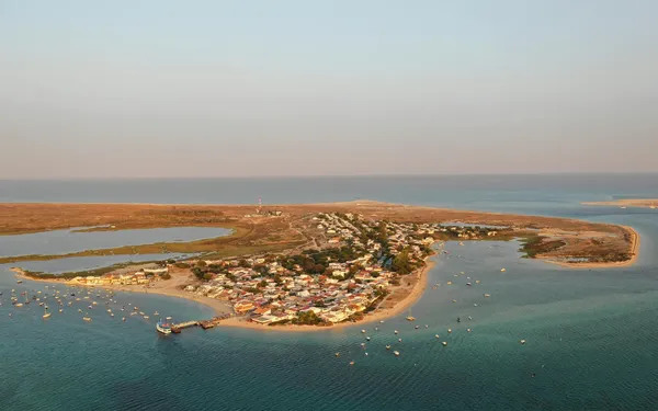 Aerial view of a small coastal town on a peninsula, surrounded by blue water and anchored boats, with a sandy beach and sparse vegetation.