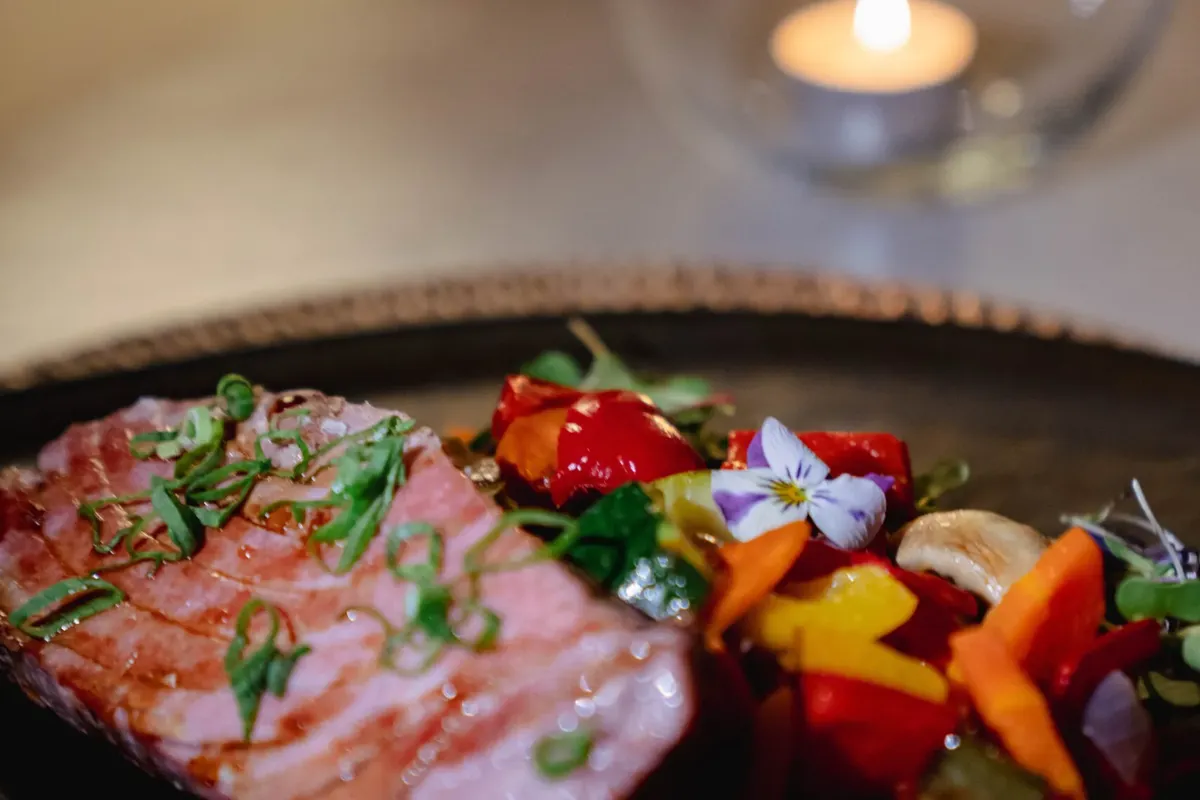 Grilled meat garnished with herbs beside vibrant mixed vegetables, served on a dark plate. Background features a glass candle holder with a lit candle.