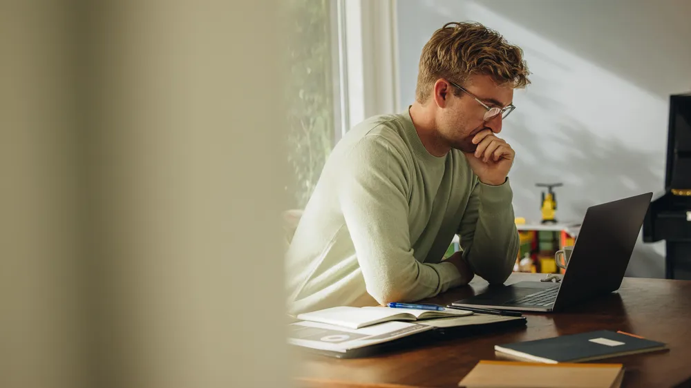Man with glasses sitting at a desk, focused on a laptop, with open notebooks and a window in the background.