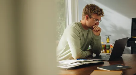 Man with glasses sitting at a desk, focused on a laptop, with open notebooks and a window in the background.