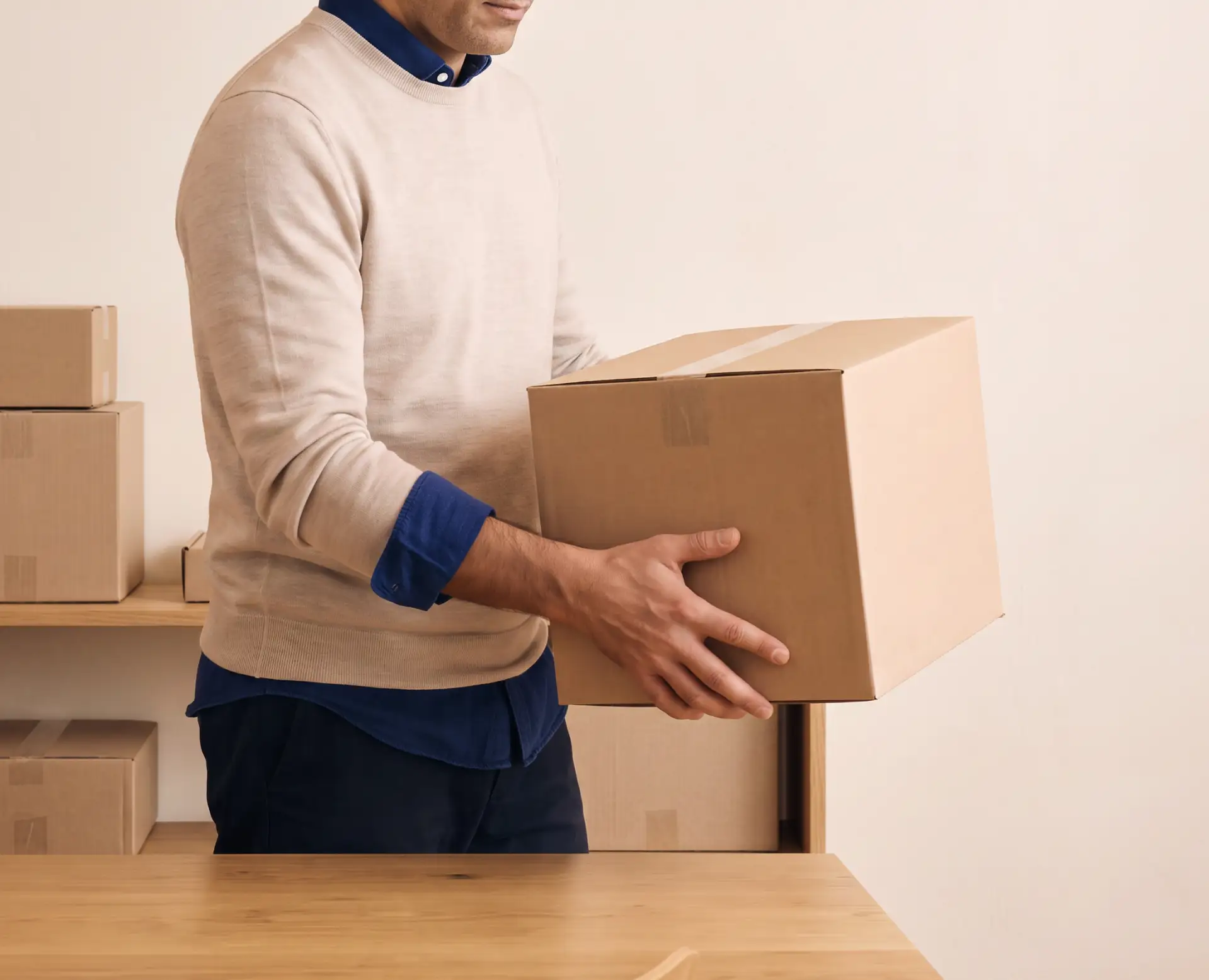 Person wearing a beige sweater holding a cardboard box, with more boxes on a wooden table and shelves in the background.