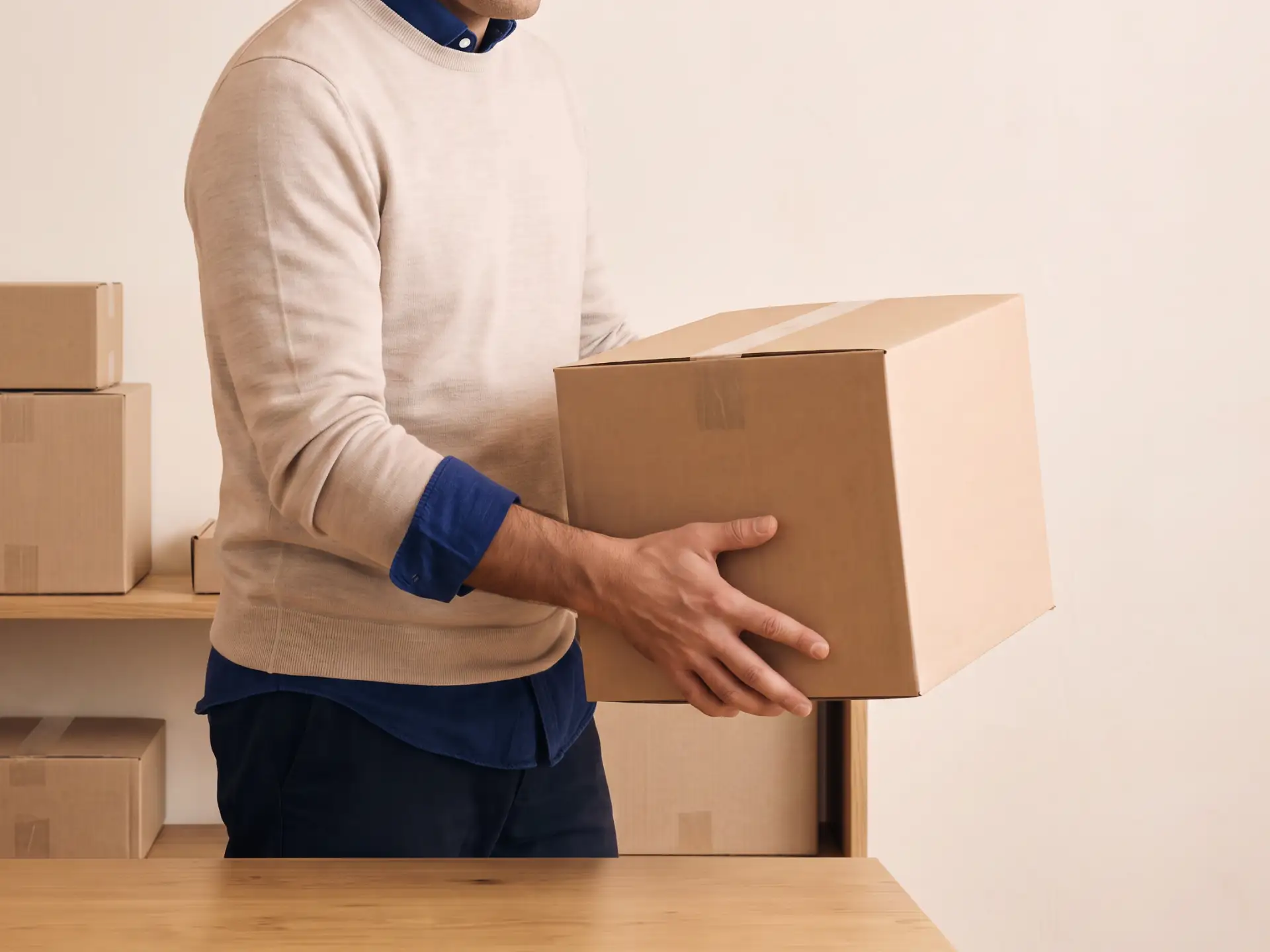 Person wearing a beige sweater holding a cardboard box, with more boxes on a wooden table and shelves in the background.
