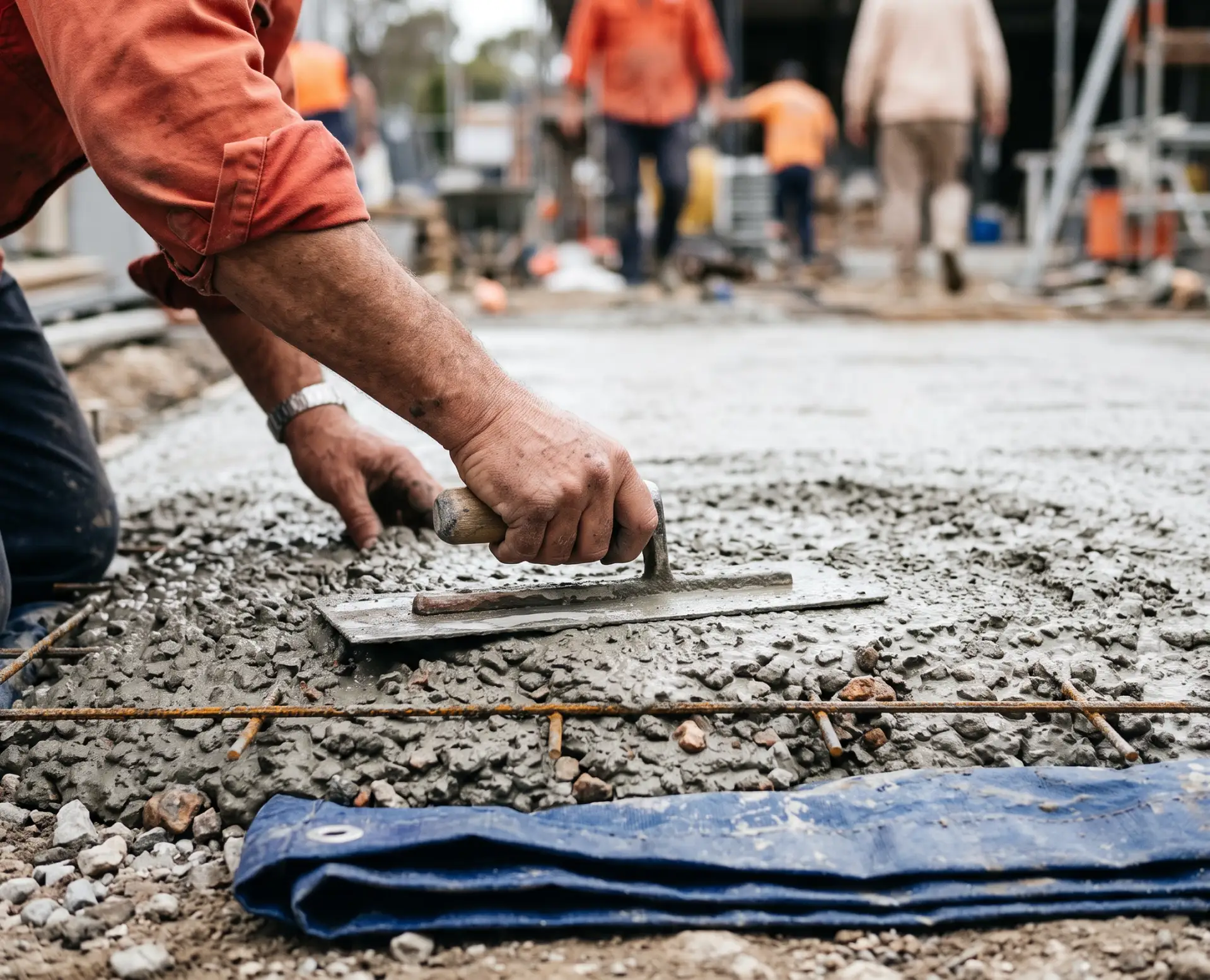 A construction worker smooths wet concrete with a trowel on a building site, with others working in the blurred background.