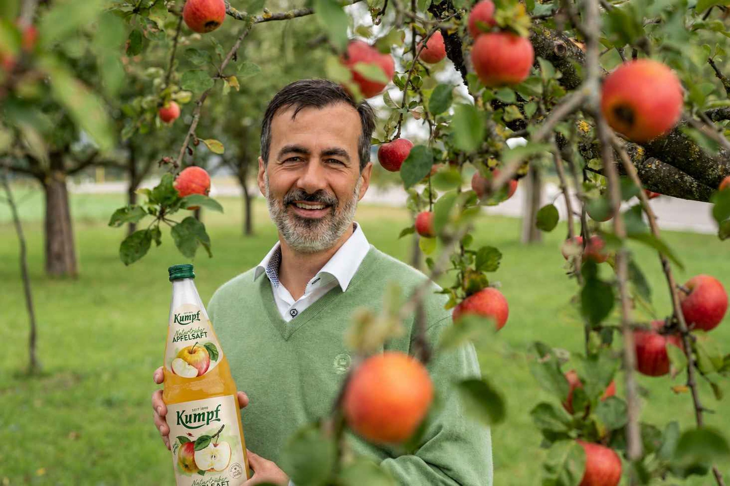 Albrecht Kumpf in einem grünen Pullover hält eine Flasche Apfelsaft in einem blühenden Apfelgarten.