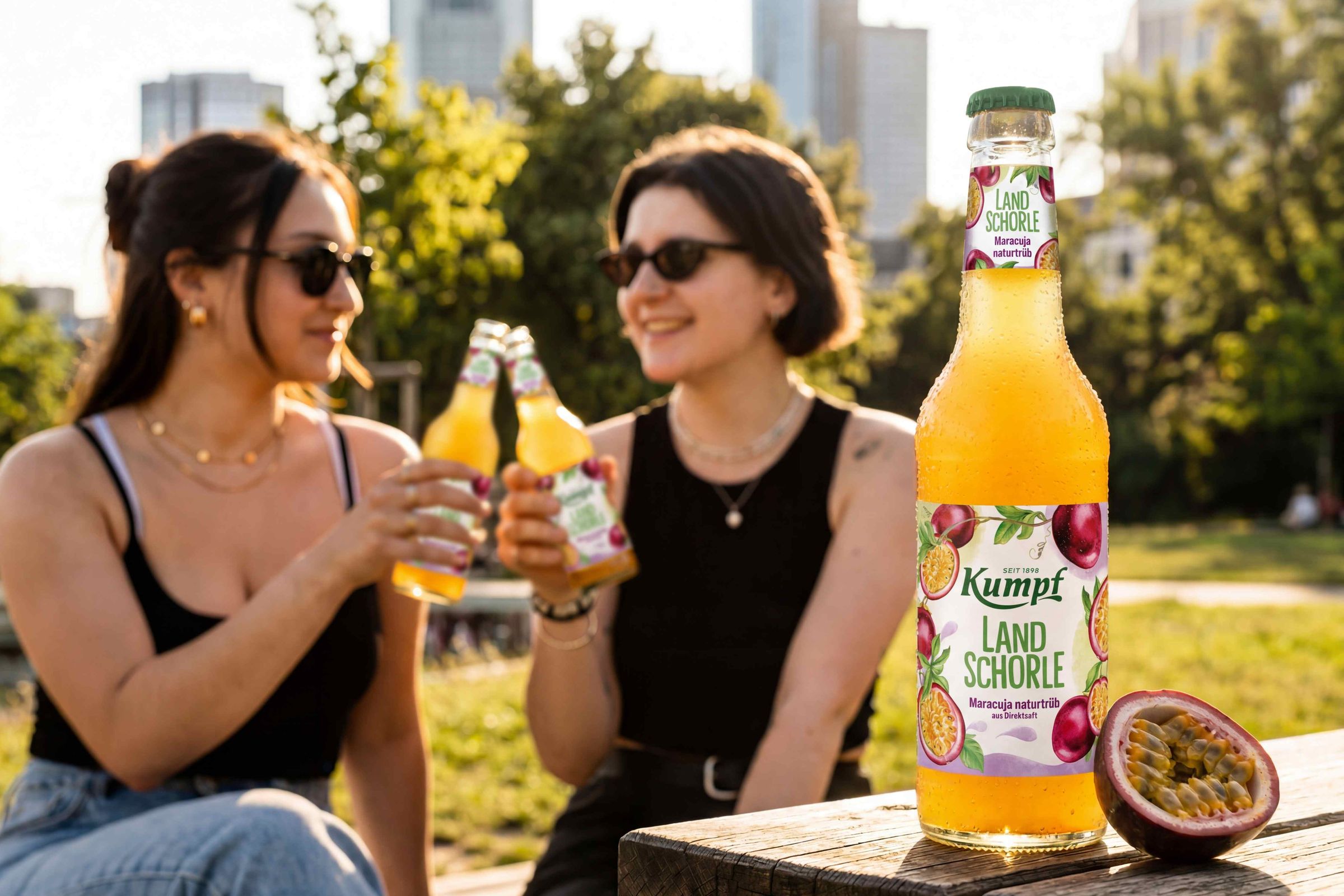 Zwei Frauen stoßen mit Getränkeflaschen an. Im Vordergrund eine Flasche Kumpf Land Schorle mit Maracuja und aufgeschnittener Frucht. Hintergrund: Parklandschaft.