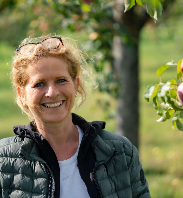 Irmgard Kumpf mit Brille auf dem Kopf steht auf einer Streuobstwiese. Im Hintergrund sind grüne Bäume und reife, rote Äpfel zu sehen.