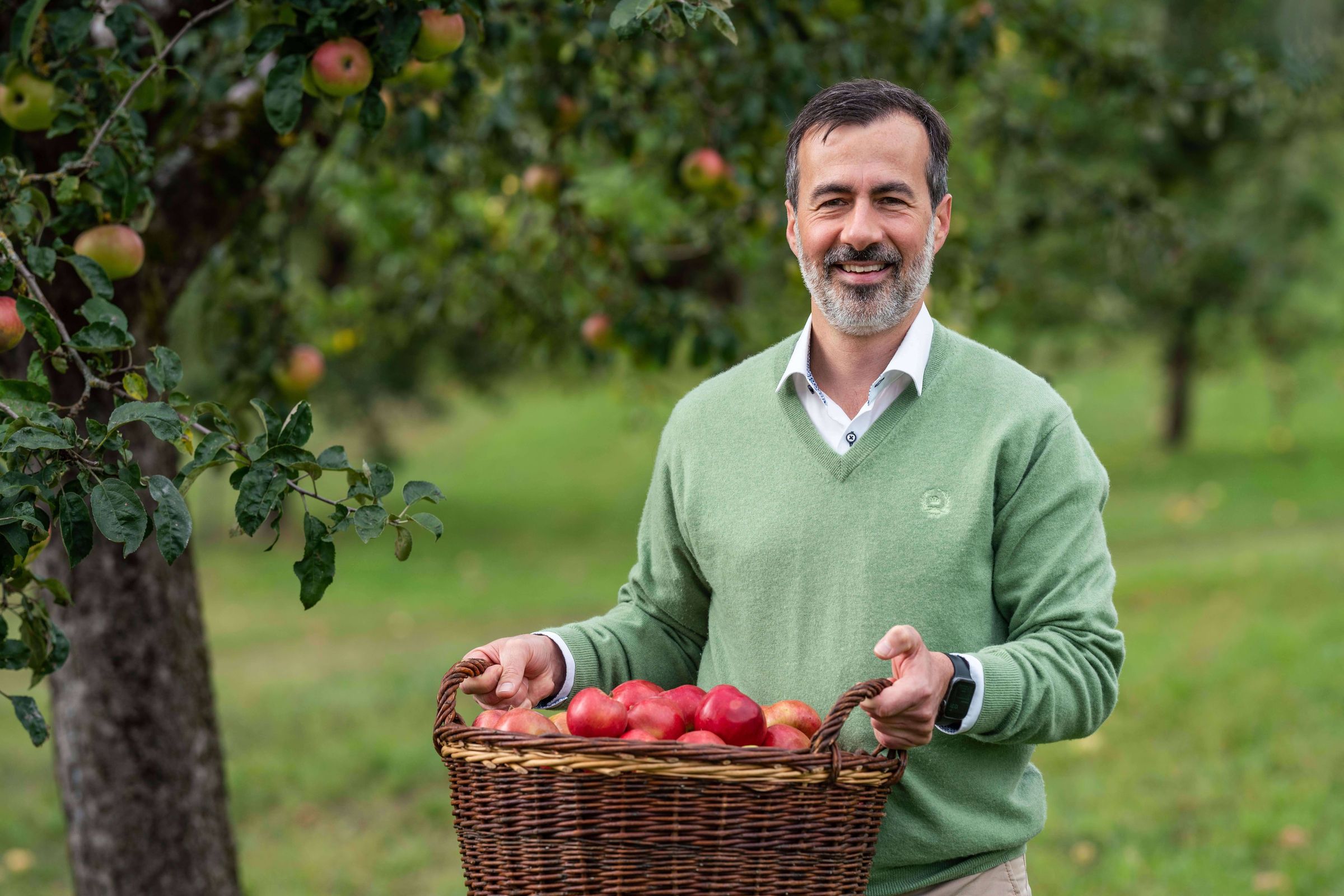 Albrecht Kumpf in einem grünen Pullover, hält einen Korb voller Äpfel auf einer Streuobstwiese. Im Hintergrund Apfelbäume.