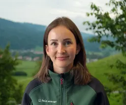 Woman in a green jacket stands outdoors with a scenic background of hills and trees.
