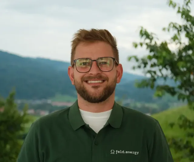 Man with glasses and beard smiling, wearing a green "feld.energy" shirt, standing outdoors with hills and trees in the background.