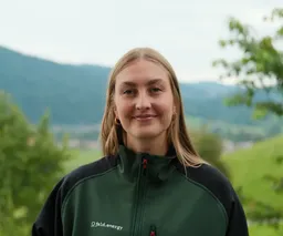 A person with long hair smiles outdoors, wearing a dark green jacket with "field.energy" text. Green hills and trees are in the background.