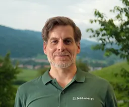 Man in a green polo shirt with "feld.energy" logo, standing outdoors with hills and trees in the background.