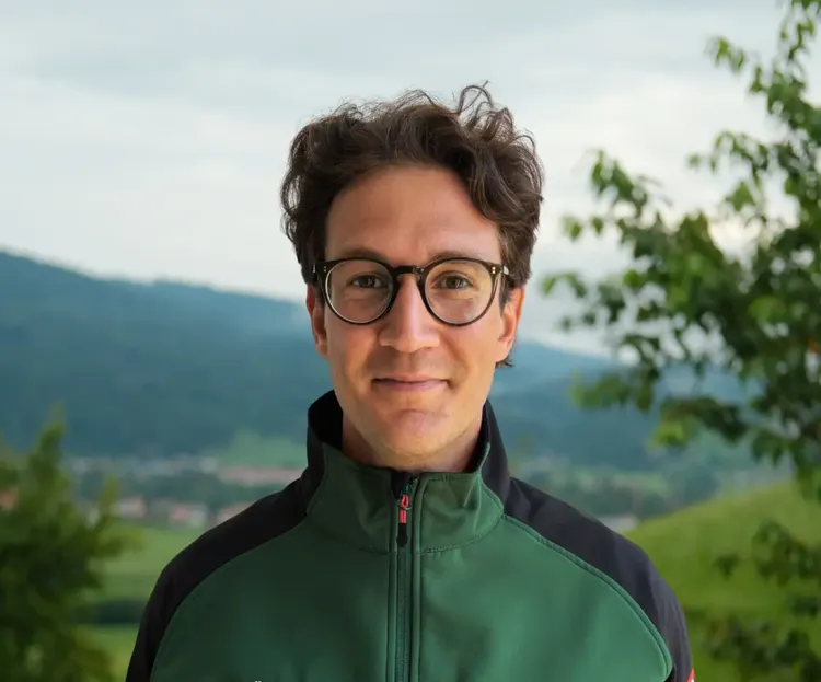 A person with glasses and curly hair stands outdoors, wearing a green jacket. A scenic background of hills and trees is visible.
