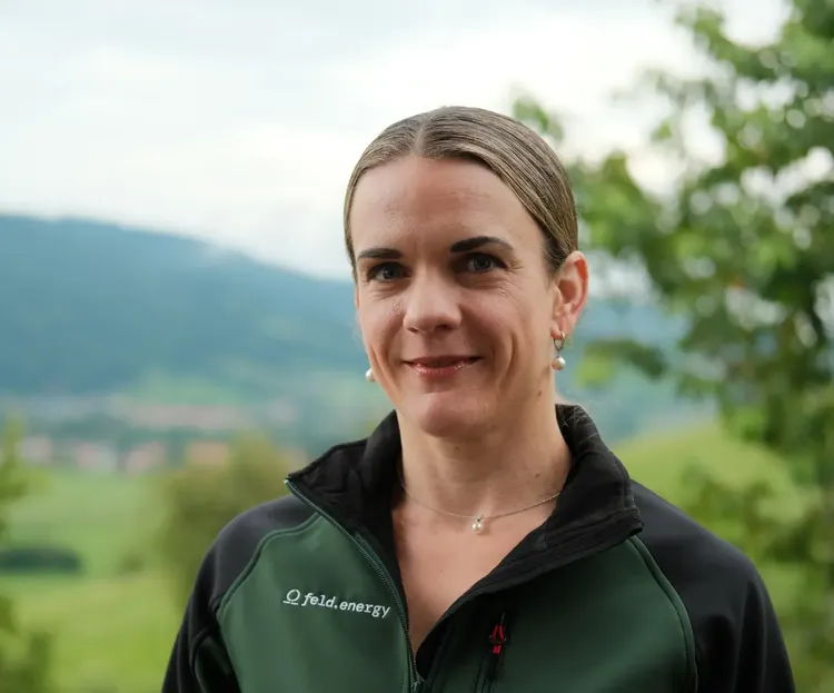A person with tied-back hair smiles outdoors, wearing a green jacket with a logo. A blurred landscape of hills and trees is in the background.