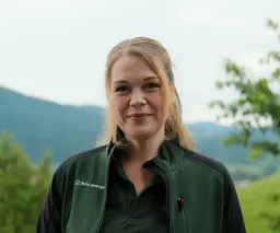 Smiling woman in green jacket stands outdoors with blurred mountains and trees in the background.