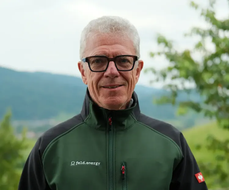 Man wearing glasses and a green jacket with "feld.energy" logo, standing outdoors with trees and hills in the background.