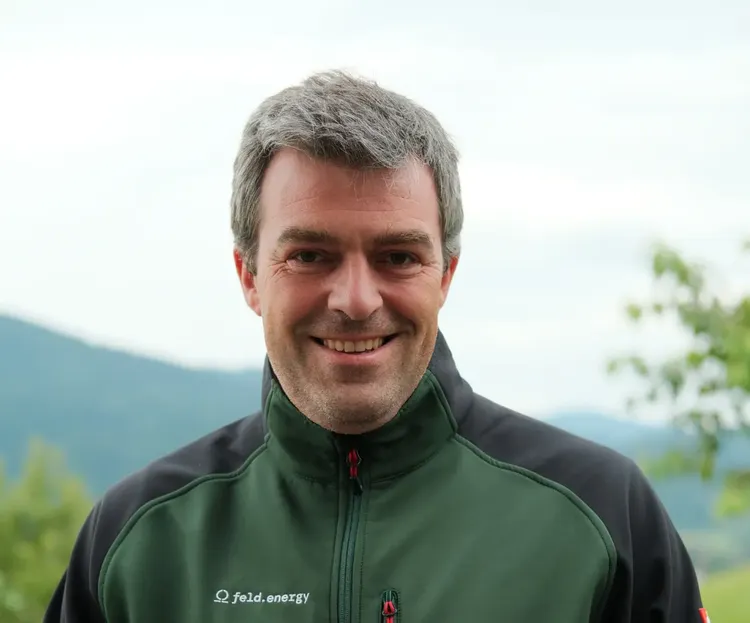 Smiling man in a green and black jacket stands outdoors with blurred hills and trees in the background.