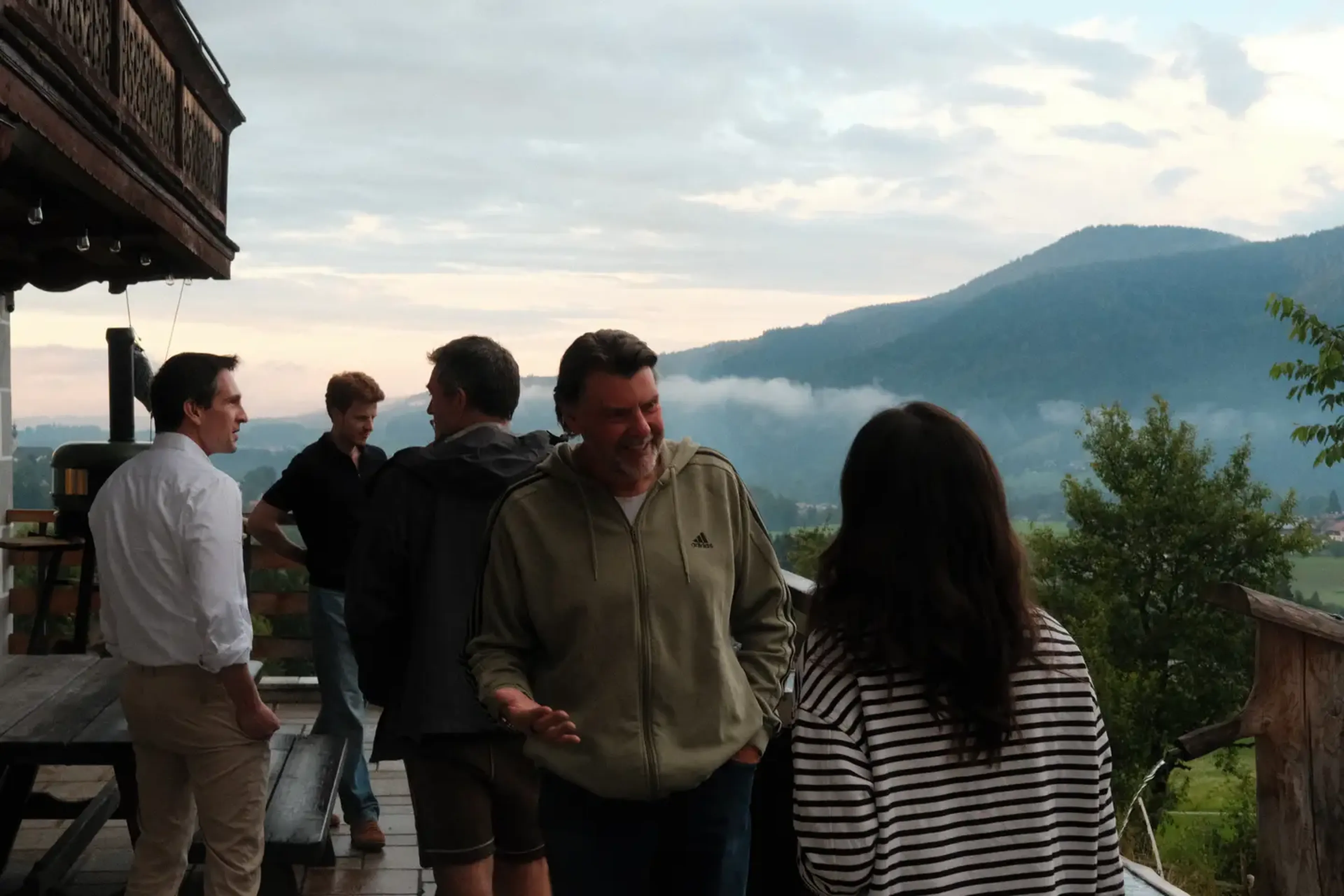 A group of people talking on a wooden deck with mountains and a cloudy sky in the background.
