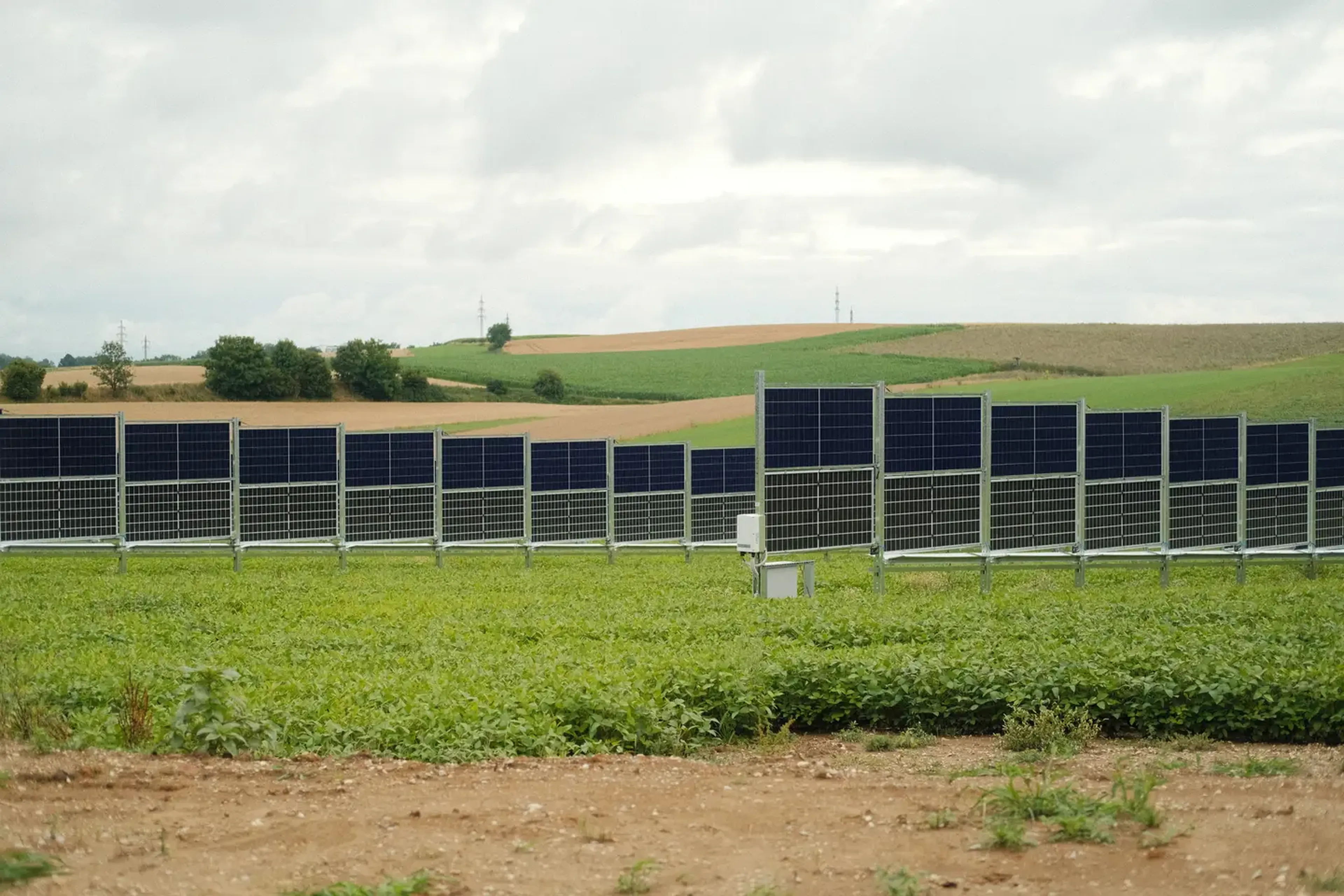 Rows of solar panels in a green field under a cloudy sky, with rolling hills in the background.