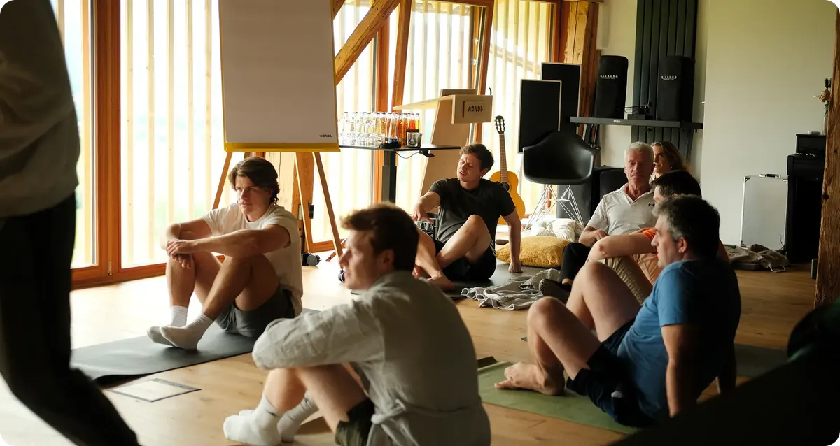 A group of people sitting on the floor in a room with large windows, engaging in a casual discussion or workshop setting.